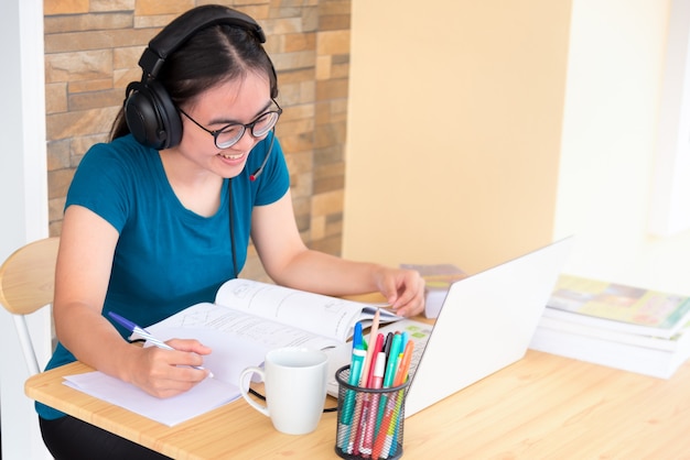 Jovem estudante adolescente asiática com fones de ouvido e óculos está sorrindo feliz olhando para o aprendizado on-line do computador portátil na escola. Aula de educação a distância da universidade videochamada em casa