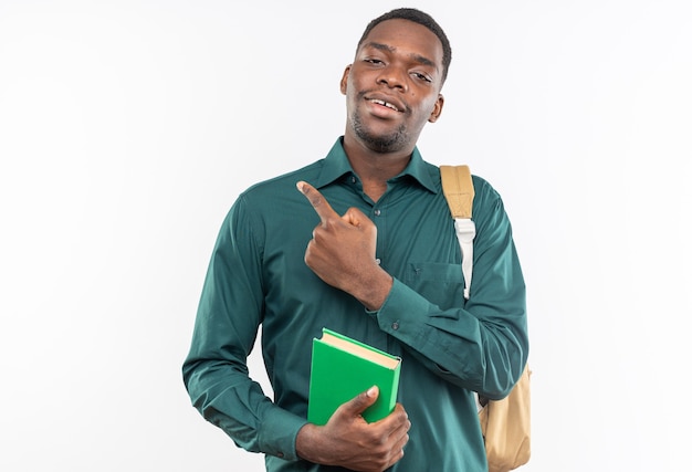 Jovem estudante afro-americana sorridente com uma mochila segurando um livro e apontando para o lado