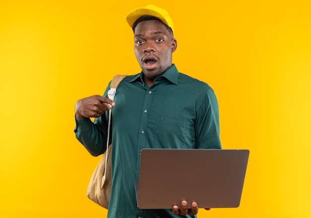 Jovem estudante afro-americana surpresa com boné e mochila segurando e apontando para laptop isolado na parede laranja com espaço de cópia