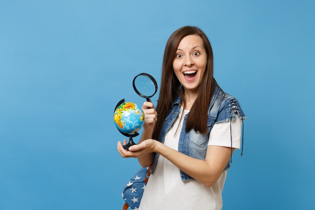 Jovem estudante animada em roupas jeans com mochila olhando no globo do mundo com lupa aprender sobre países isolados sobre fundo azul. educação na faculdade universitária do ensino médio.