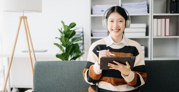 Jovem estudante asiática atraente e feliz estudando na biblioteca da faculdade sentada no sofá usando um computador portátil, tablet e fones de ouvido conversando por vídeo