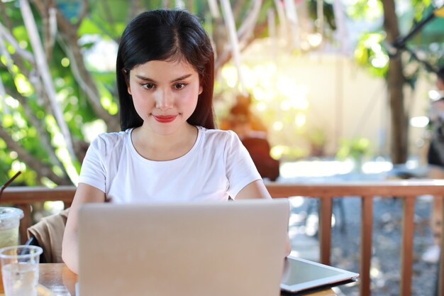 Jovem estudante asiática, inteligente e ativa, sentada e usando o computador, anotando para estudar online com uma cara sorridente e feliz (novo conceito normal)