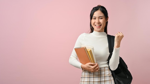 Jovem estudante asiático segurando livros com sinal sim em fundo rosa isolado