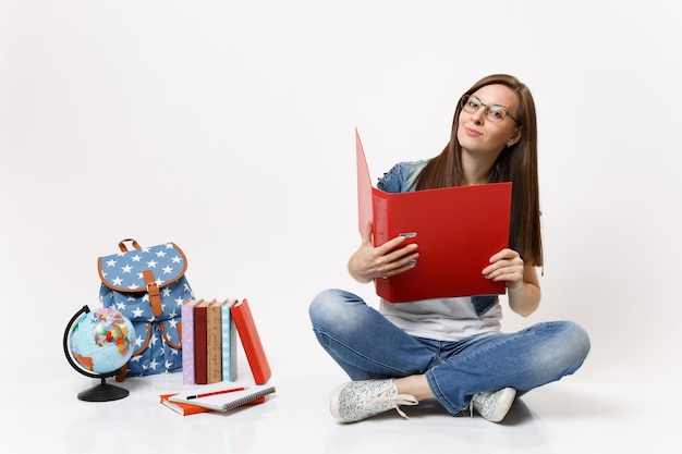 Jovem estudante atraente de óculos segurando uma pasta vermelha para documentos, sentada perto da mochila globo, livros escolares isolados