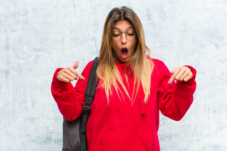 Foto jovem estudante bonita se sentindo chocada, boquiaberta e espantada, olhando e apontando para baixo com descrença e surpresa