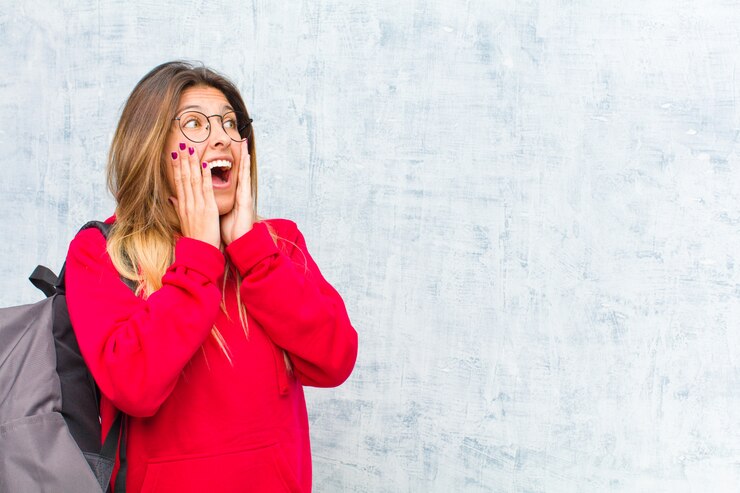 Foto jovem estudante bonita se sentindo feliz, animada e surpresa, olhando para o lado com as duas mãos no rosto