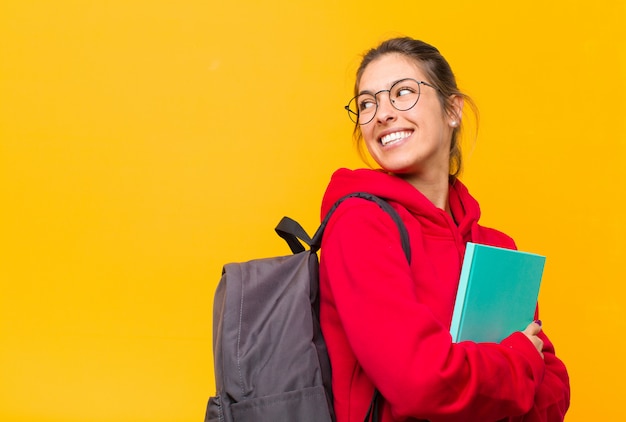 Jovem estudante bonita sorrindo alegremente, sentindo-se feliz, satisfeito e relaxado, com os braços cruzados e olhando para o lado