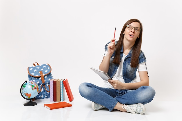 Jovem estudante casual inteligente, lembrando-se de pensar em pensar olhando para cima, apontando o lápis para cima perto da mochila globo, livros escolares isolados