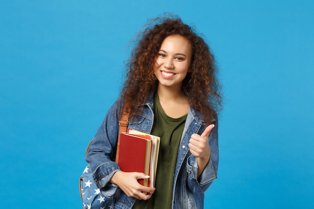 Jovem estudante com roupas jeans e mochila segurando livros isolados na parede azul