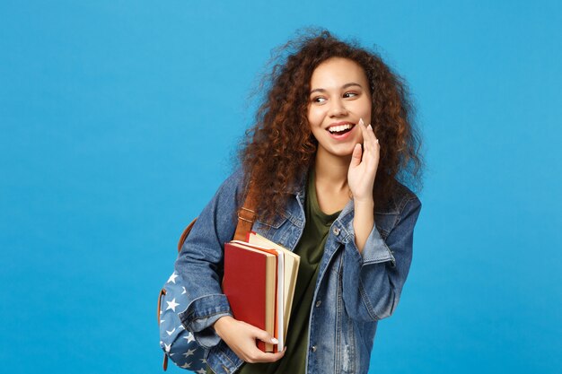 Jovem estudante com roupas jeans e mochila segurando livros isolados na parede azul