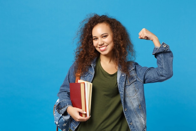 Jovem estudante com roupas jeans e mochila segurando livros isolados na parede azul