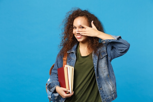 Jovem estudante com roupas jeans e mochila segurando livros isolados na parede azul