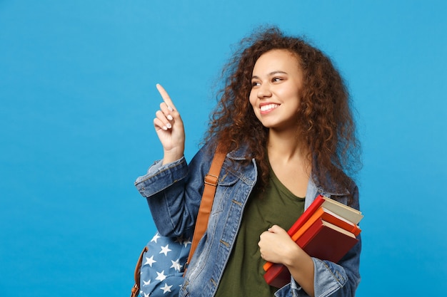 Jovem estudante com roupas jeans e mochila segurando livros isolados na parede azul