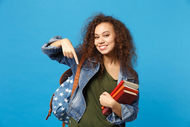 Jovem estudante com roupas jeans e mochila segurando livros isolados na parede azul