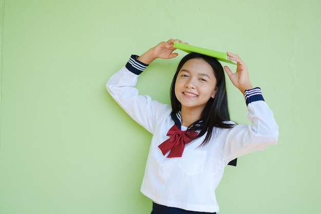 Jovem estudante de retrato na escola uniforme com um livro verde na parede verde. Adolescente asiática