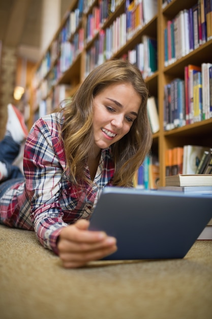 Jovem estudante deitado no chão da biblioteca usando um tablet