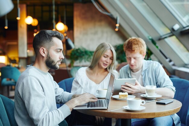 Jovem estudante em trajes casuais sentado à mesa em um café em frente ao laptop e rede no fundo de seus amigos usando o touchpad