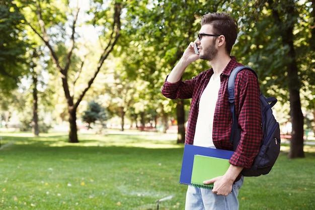 Jovem estudante falando no smartphone, segurando notebooks e mochila atrás dos ombros no parque. tecnologia, comunicação, educação e conceito de trabalho remoto, espaço de cópia