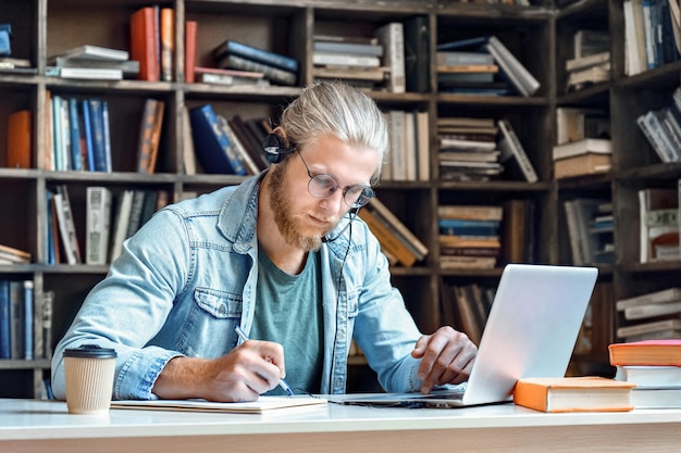 Jovem estudante focado usa fone de ouvido fazer anotações e estudar on-line na biblioteca ouvir palestra curso de internet treinamento on-line aprendizagem de língua estrangeira educação a distância escrever informações no caderno