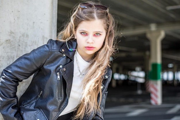 Jovem estudante loira de jaqueta de couro preta posando no estacionamento