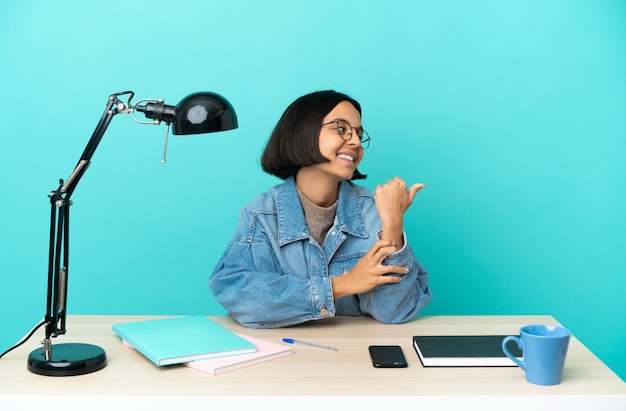 Jovem estudante mestiça estudando sobre uma mesa apontando para o lado para apresentar um produto