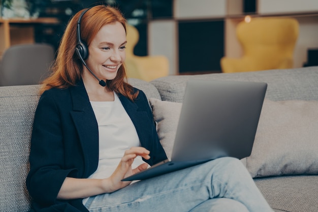 Jovem estudante ruiva positiva tendo webinar on-line via computador laptop e fones de ouvido enquanto está sentada no sofá em uma cafeteria, estudando remotamente em um local público usando a tecnologia