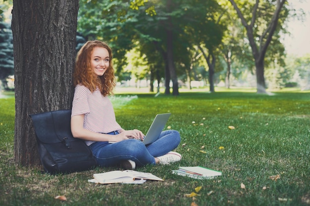 Jovem estudante ruiva sorridente na digitação casual no laptop, sentado sob uma árvore no parque. Conceito de educação