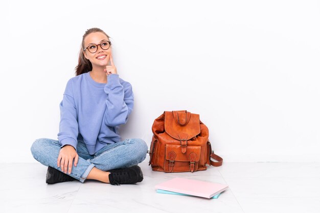 Foto jovem estudante sentada no chão, isolada no fundo branco, pensando em uma ideia enquanto olha para cima