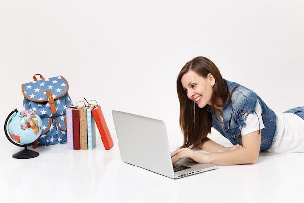Jovem estudante sorridente em roupas jeans, trabalhando em um computador laptop, deitada perto da mochila globo e livros escolares isolados