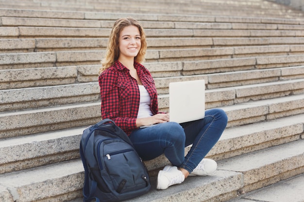 Jovem estudante sorridente sentada nas escadas usando laptop ao ar livre durante as férias descansando no campus universitário. tecnologia, educação e conceito de trabalho remoto, copie o espaço