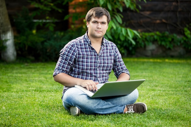 Jovem estudante sorridente, sentado na grama do parque e digitando no laptop