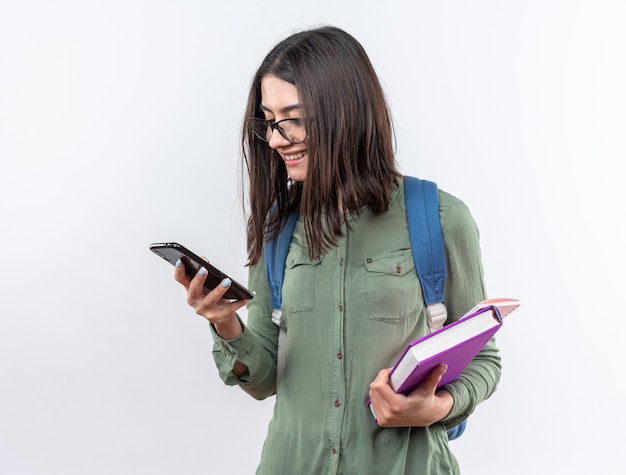 Jovem estudante sorridente usando óculos com uma mochila segurando livros e olhando para o telefone na mão