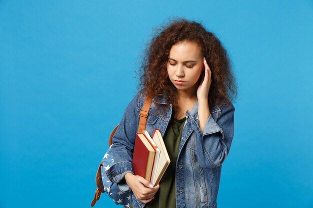 Jovem estudante triste com roupas jeans e mochila segurando livros isolados na parede azul