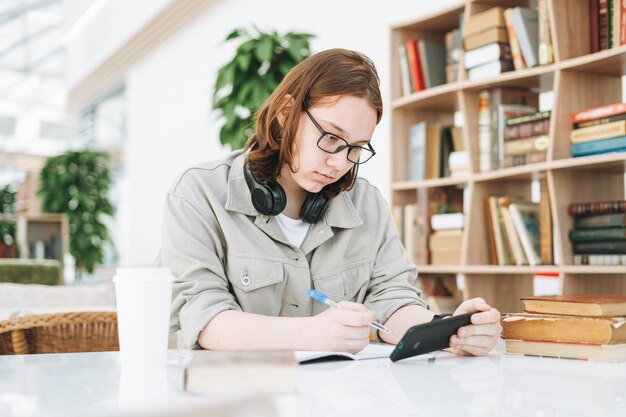 Jovem estudante universitária de óculos fazendo lição de casa com telefone celular na biblioteca moderna