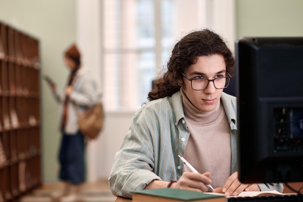 Jovem estudante usando óculos olhando para o computador na biblioteca