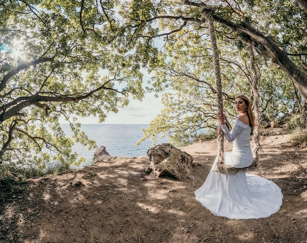 Jovem europeia bonita em vestido de noiva branco posando em balanço em árvores da floresta no fundo da praia do mar do oceano Idéias exóticas para fotografia de casamento