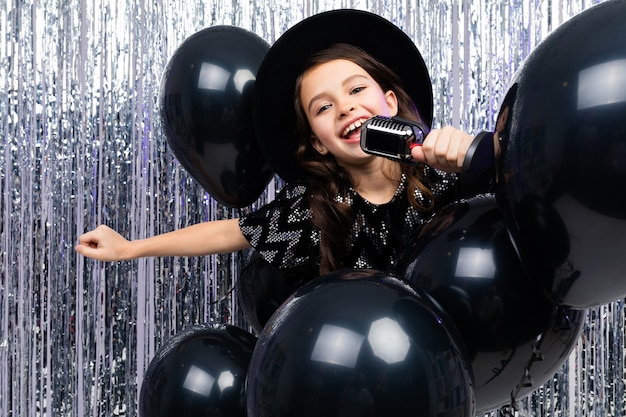 jovem europeia cantando com um microfone em uma festa contra um de balões pretos e enfeites de Natal