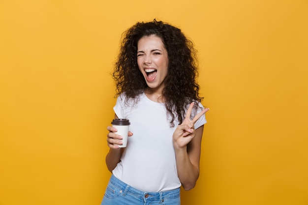 jovem europeia de 20 anos com cabelo encaracolado, sorrindo e segurando um café para viagem em copo de papel isolado em amarelo