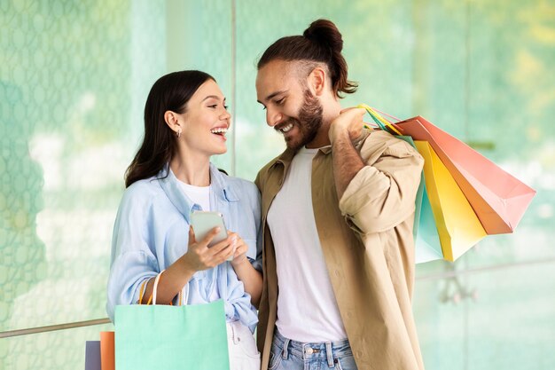 Foto jovem europeia sorridente mostra smartphone para homem com sacolas, recomenda aplicativo, aproveite as compras