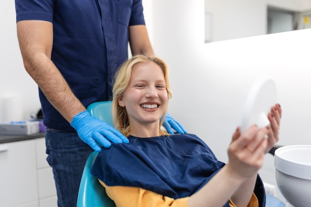 Jovem europeia sorrindo enquanto olha para o espelho na clínica odontológica Foto de uma jovem verificando seus resultados no consultório do dentista