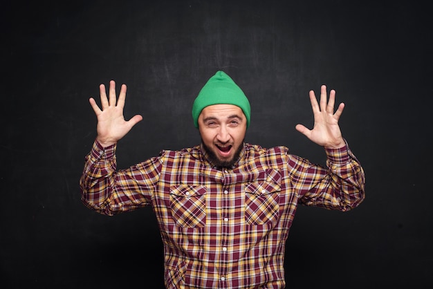 Foto jovem europeu com barba com chapéu de malha verde, parece surpreso e perplexo. mostrando os dedos para cima e para o lado direito. fundo preto, espaço de cópia em branco para texto ou anúncio