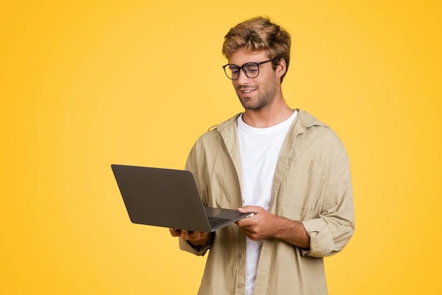 Jovem europeu de óculos segura laptop em pano de fundo de estúdio amarelo