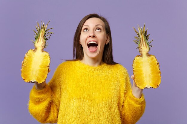 Jovem excitada olhando para cima mantendo a boca aberta segurando metades de frutas frescas de abacaxi maduro isoladas em fundo pastel violeta. Estilo de vida vívido de pessoas, relaxe o conceito de férias. Mock up espaço de cópia.