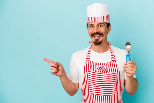 Jovem fabricante caucasiano segurando uma colher isolada no fundo azul, sorrindo e apontando de lado, mostrando algo no espaço em branco.