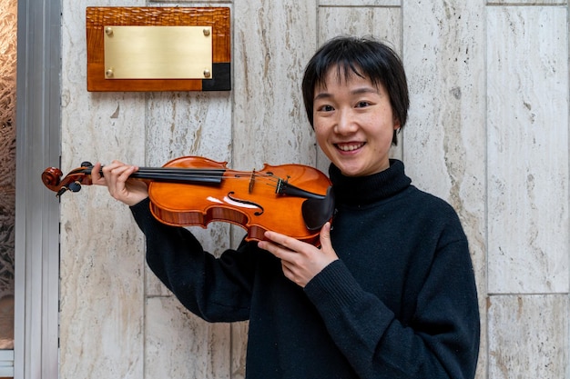 Foto jovem fabricante de violinos chinesa mostrando seu violino recém-feito ao ar livre na frente de sua oficina