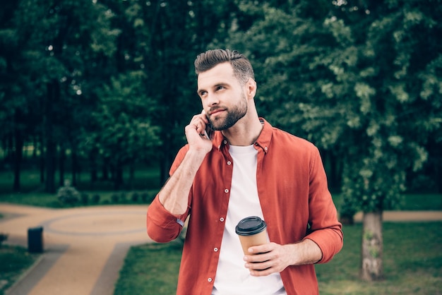 Jovem falando no smartphone e segurando café para viagem em pé no parque