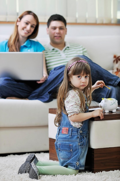 jovem família feliz na sala de estar brilhante e moderna, colocando dinheiro no cofrinho e trabalhando no computador portátil em finanças domésticas