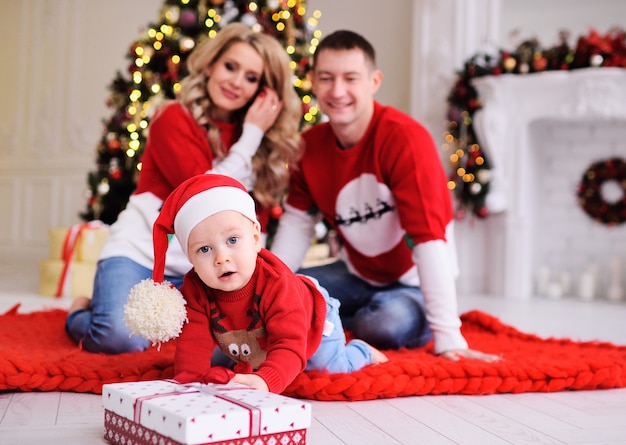 Jovem família-mãe, pai e filho pequeno na árvore de Natal. Um menino de chapéu vermelho do Papai Noel rasteja para um presente de Natal.