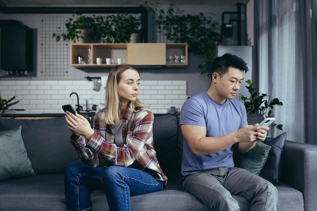 Foto jovem família multirracial brigando sentada no sofá na sala, homem e mulher ao telefone, viraram em direções diferentes