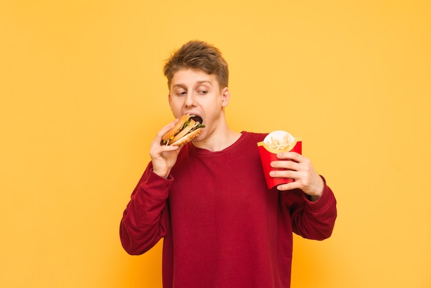 Jovem faminto em roupas casuais com batatas fritas nas mãos e morde um hambúrguer em um amarelo